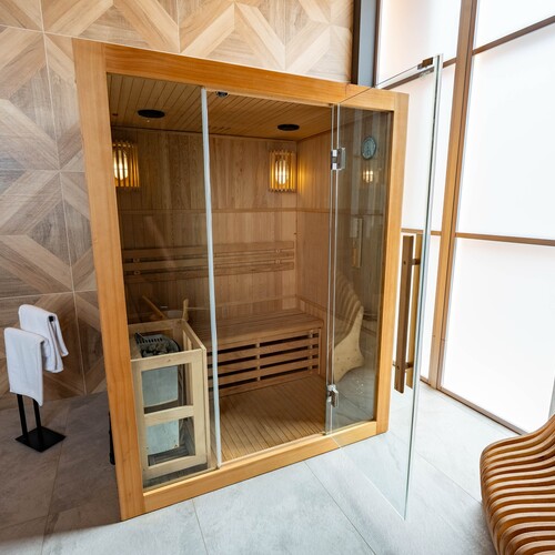 Private wooden sauna with glass door, benches, stone heater, warm lighting and towel rack.