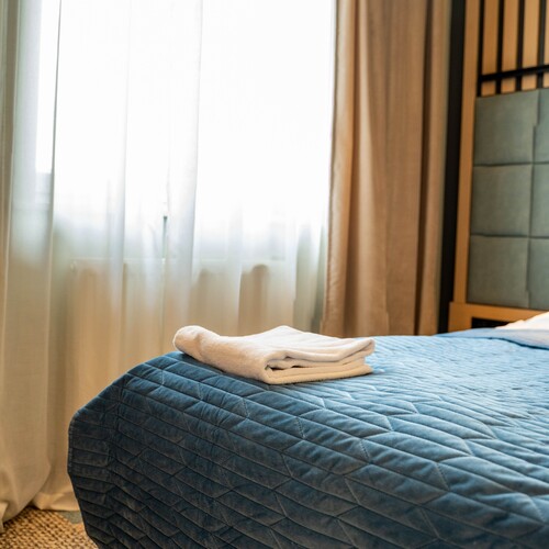 Quilted blue bedspread with neatly folded white towels on a made bed by sheer curtains and soft light.