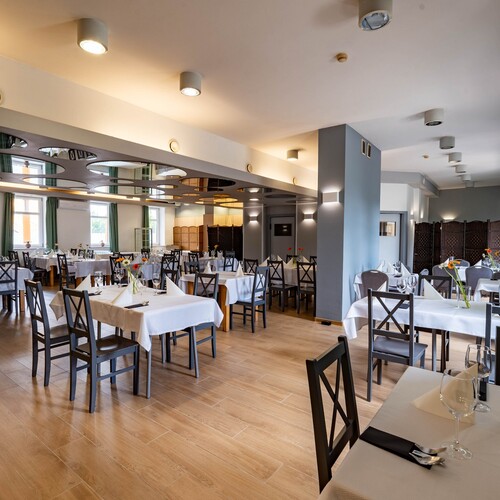 Spacious hotel restaurant with white tablecloths, place settings and wine glasses, wooden chairs, green curtains and round ceiling panels