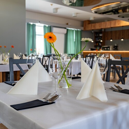Hotel restaurant with white tablecloths, folded napkins, wine glasses and gerberas in vases; modern wood bar and green curtains.