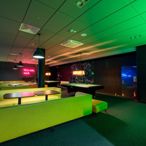 Hotel games room with 'drink eat play' neon, green sofas, pool tables, bowling balls and colorful lighting.