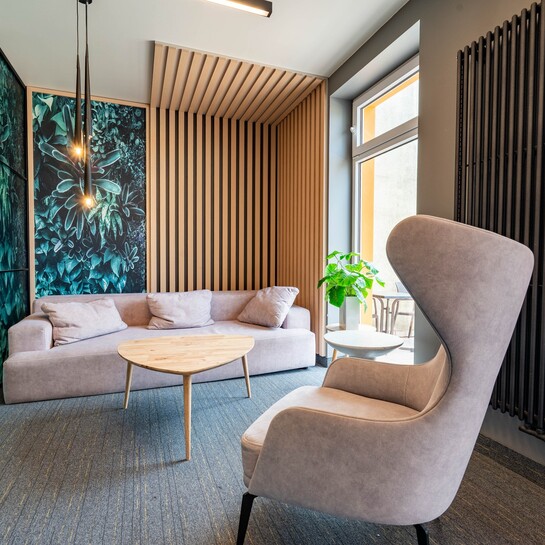 Nowoczesny hotelowy salon z miękką sofą i fotelem, drewnianą ścianą z lamelami, roślinnym muralem, lampami i stolikami.