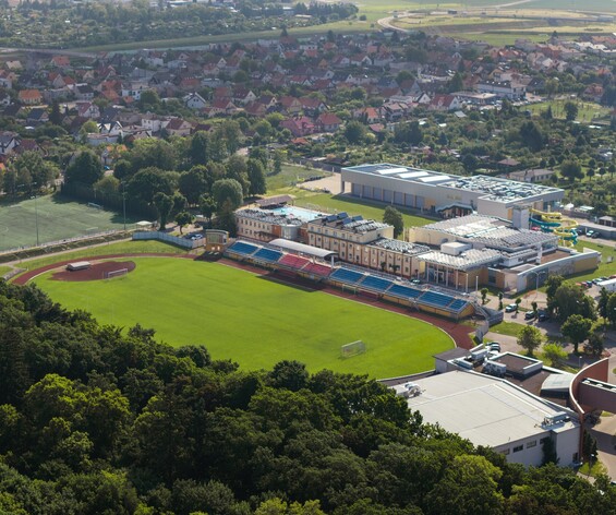 Centrum rekreacyjne z odkrytym basenem i żółtymi zjeżdżalniami obok stadionu z trybunami, w otoczeniu drzew i domów.