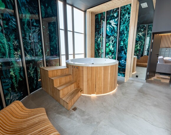Prywatna strefa spa z okrągłym drewnianym jacuzzi, schodkami i podświetleniem, roślinne szklane panele, wysokie matowe przeszklenia i leżanka.