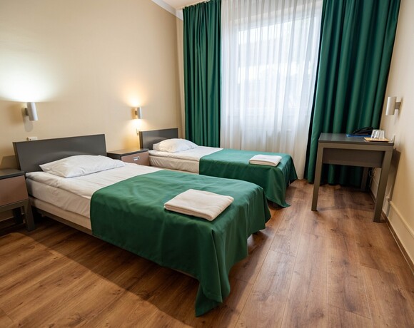 Pokój z dwoma pojedynczymi łóżkami, zielonymi narzutami, stolikami nocnymi i lampkami, biurko z telefonem i szklankami.