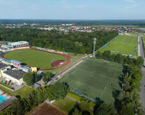 Główny stadion trawiasty z czerwoną bieżnią, boiska treningowe, zalesiony park i zabudowa mieszkaniowa w tle.