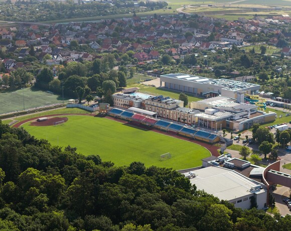 Stadion z zielonym boiskiem i bieżnią, trybuny oraz przyległy kompleks z basenem i żółtymi zjeżdżalniami, domy i zieleń w tle