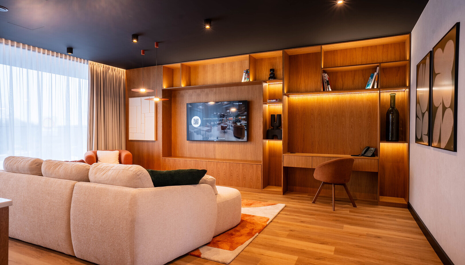 Przestronny apartament z jasną, zaokrągloną sofą, ścianą z drewnianymi półkami i TV, biurko, duże zasłonięte okno i ciepłe oświetlenie.