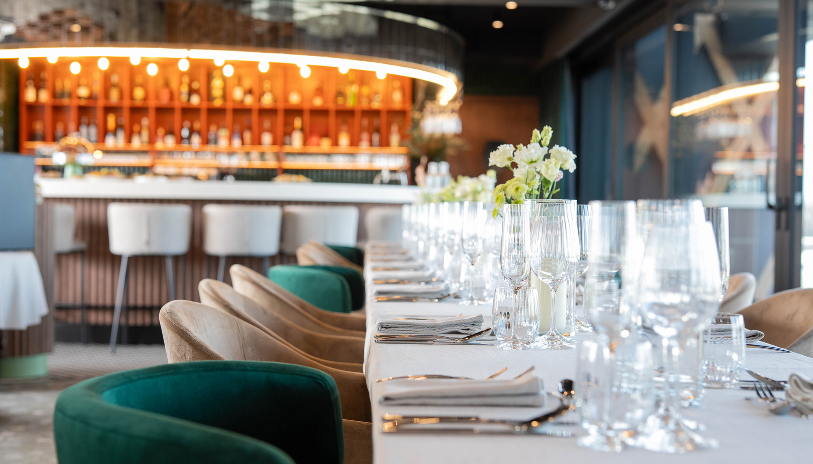 Długo nakryty stół w eleganckiej restauracji hotelowej, kryształowe kieliszki, serwetki, zielone fotele i podświetlony bar