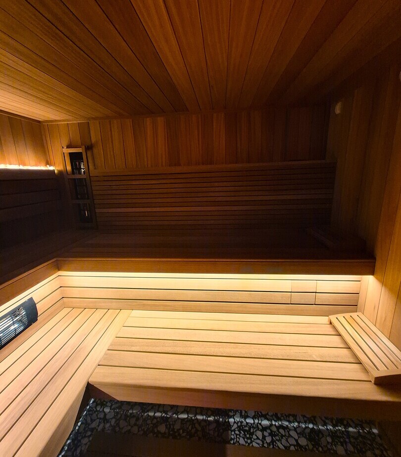 Sauna hotelowa z drewnianymi, wielopoziomowymi ławami, ciepłym ukrytym oświetleniem LED i piecem z kamieniami