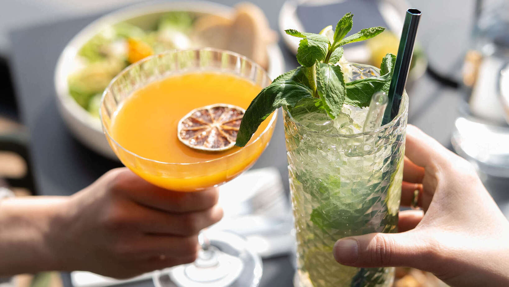 Dwa toasty: pomarańczowy koktajl z suszoną cytryną i orzeźwiający miętowy drink z kruszonym lodem, w tle talerze sałatek.