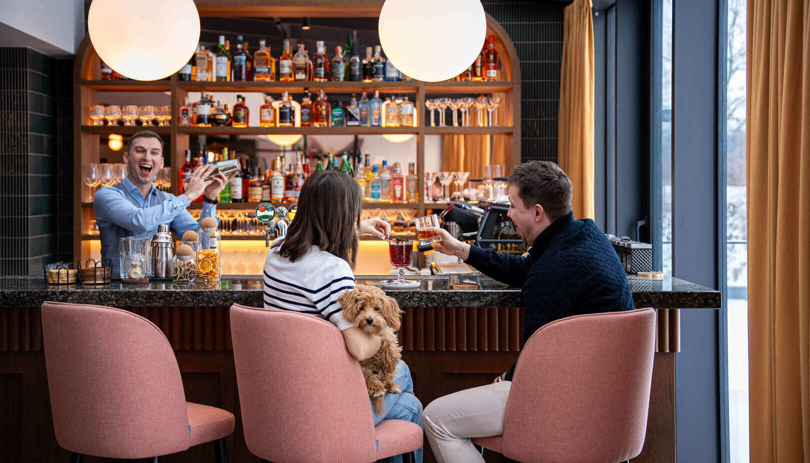 Przytulny bar hotelowy z podświetloną półką alkoholi, barman miesza koktajl, para z psem stukają kieliszkami.