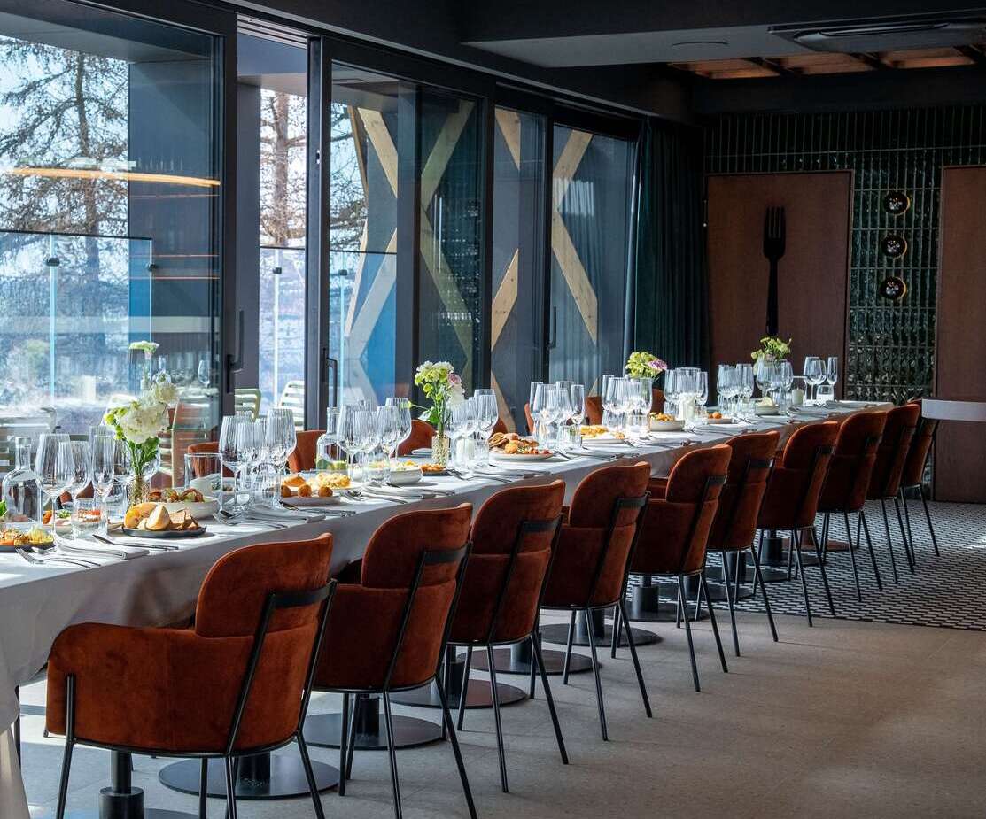 Elegancka sala restauracyjna z długim stołem nakrytym białym obrusem, brązowe fotele, szkło i kwiatowe dekoracje, widok przez duże okna.