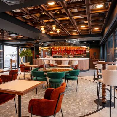 Nowoczesny hotelowy bar z podświetloną półokrągłą ladą, kasetonowym drewnianym sufitem, zielonymi i rdzawymi welurowymi fotelami i panoramicznymi oknami.