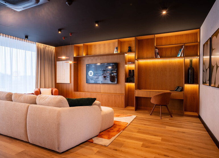 Przestronny apartament z jasną, zaokrągloną sofą, ścianą z drewnianymi półkami i TV, biurko, duże zasłonięte okno i ciepłe oświetlenie.