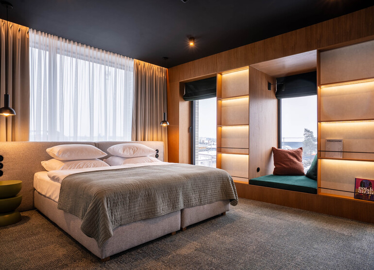 Elegancki pokój hotelowy z dużym łóżkiem, wiszącymi lampami, siedziskiem przy oknie i podświetlanymi drewnianymi półkami
