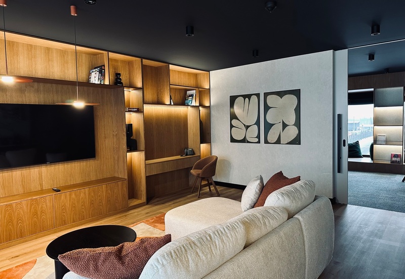 Luksusowy salon suite z jasną sofą, telewizorem, zabudowanymi drewnianymi półkami, biurkiem, nastrojowym oświetleniem i widokiem na nabrzeże.