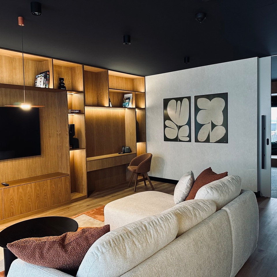 Luksusowy salon suite z jasną sofą, telewizorem, zabudowanymi drewnianymi półkami, biurkiem, nastrojowym oświetleniem i widokiem na nabrzeże.
