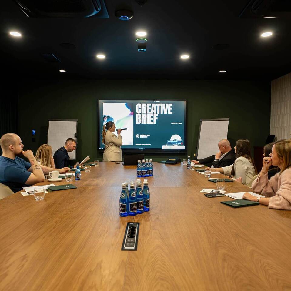Nowoczesna sala konferencyjna z długim drewnianym stołem, prezenterka przy ekranie z napisem Creative Brief, uczestnicy i woda.