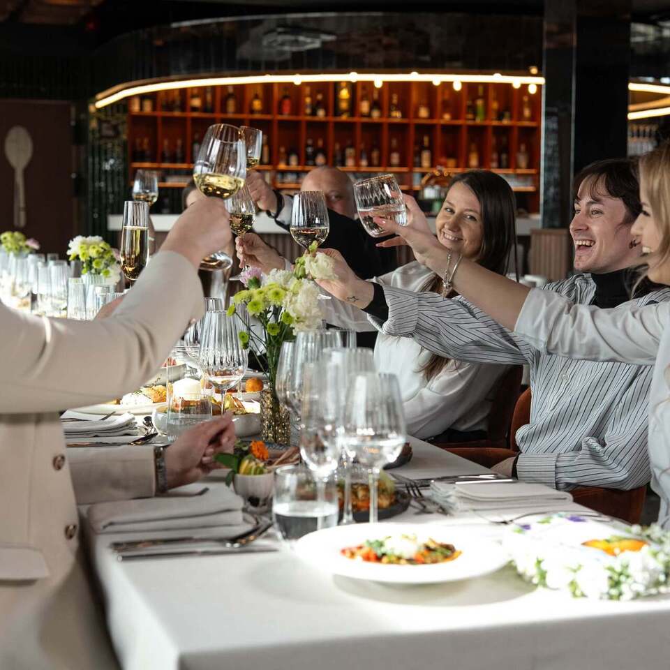 Grupa gości w eleganckiej restauracji wznosi kieliszki przy udekorowanym stole; bar, kwiaty i podane dania w tle.