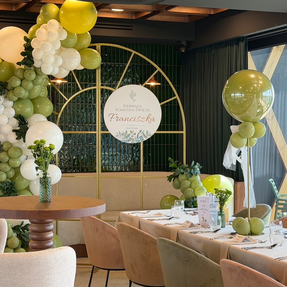 Elegancka sala z zielono-białym łukiem balonów, złotą ramą i nakrytym stołem przygotowanym na przyjęcie komunijne.
