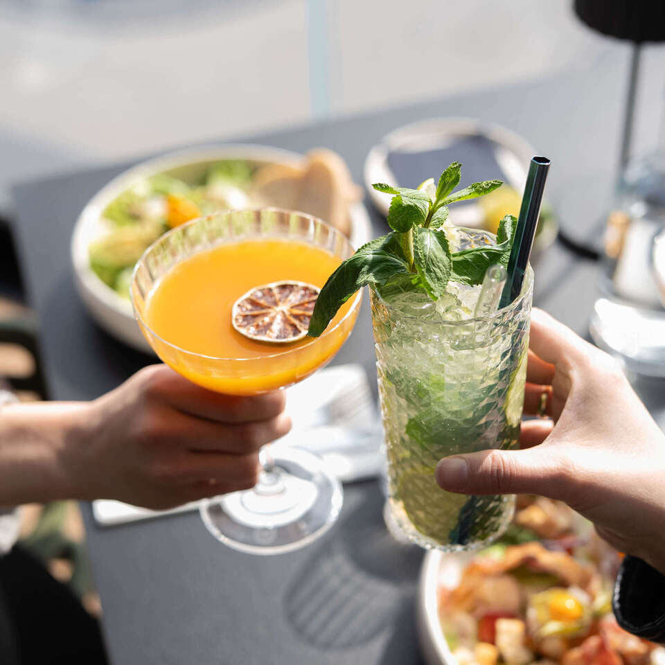 Dwa toasty: pomarańczowy koktajl z suszoną cytryną i orzeźwiający miętowy drink z kruszonym lodem, w tle talerze sałatek.