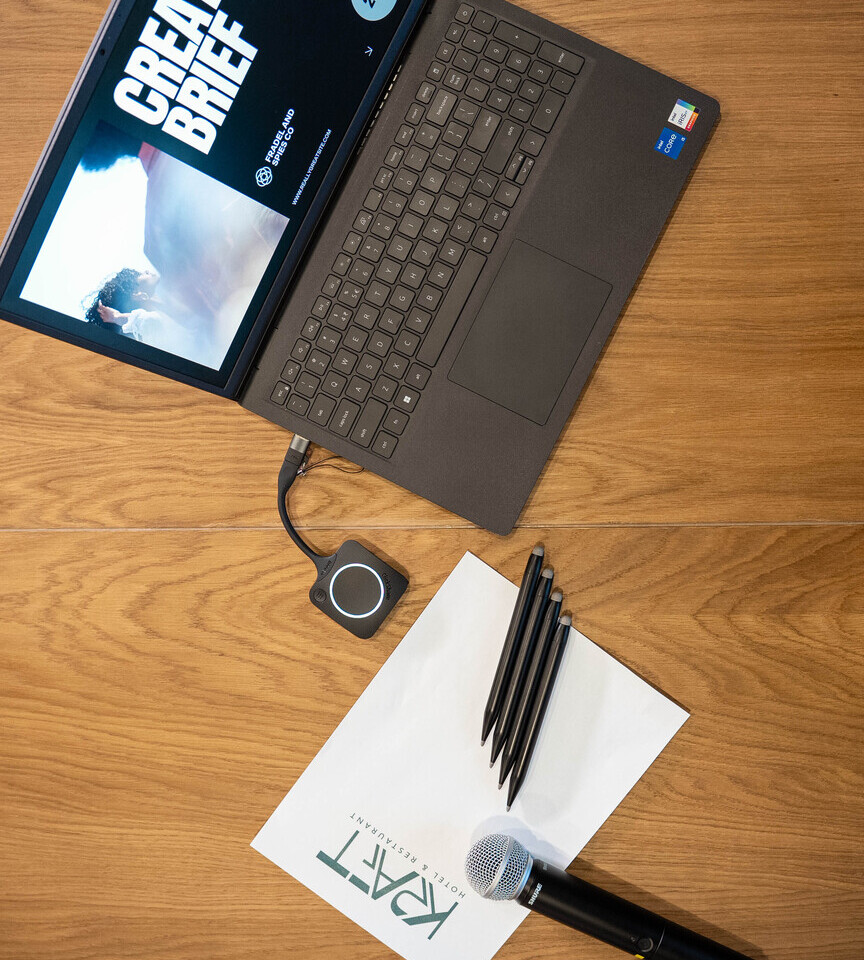 Laptop z pokazem CREATIVE BRIEF, bezprzewodowy prezenter, mikrofon, długopisy i papier z logo hotelu na drewnianym stole