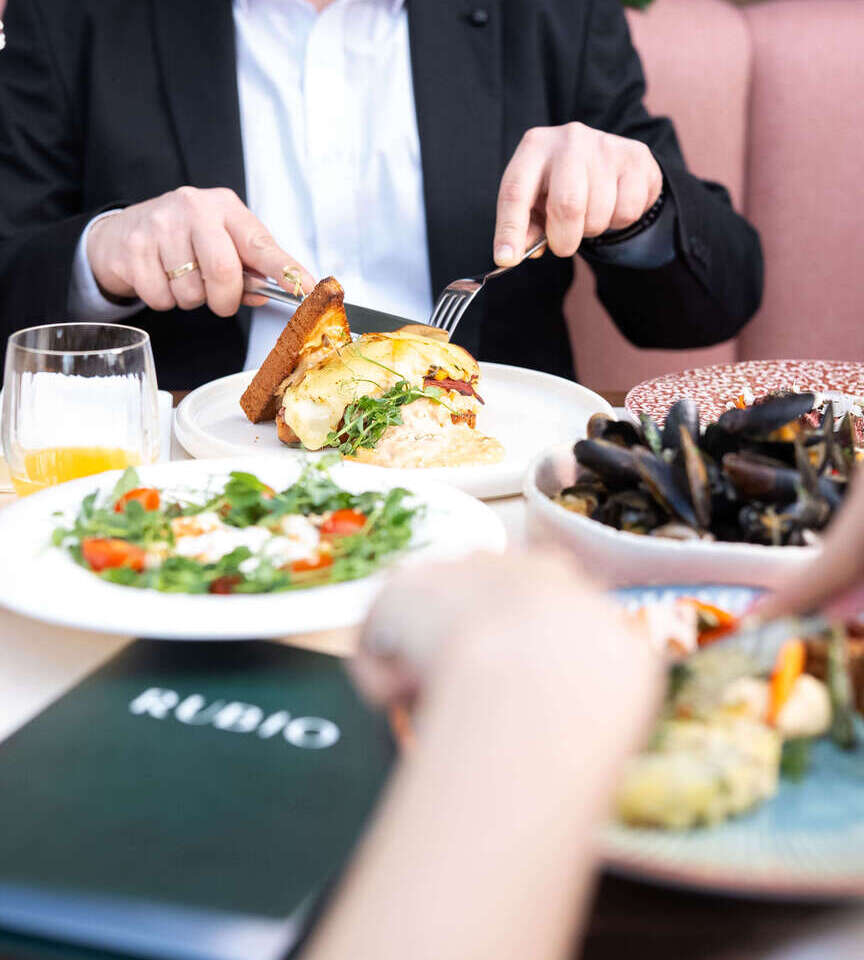 Elegancki gość w garniturze kroi jajka benedyktyńskie na toście z sosem holenderskim; obok sałatka, miska małży i menu restauracji