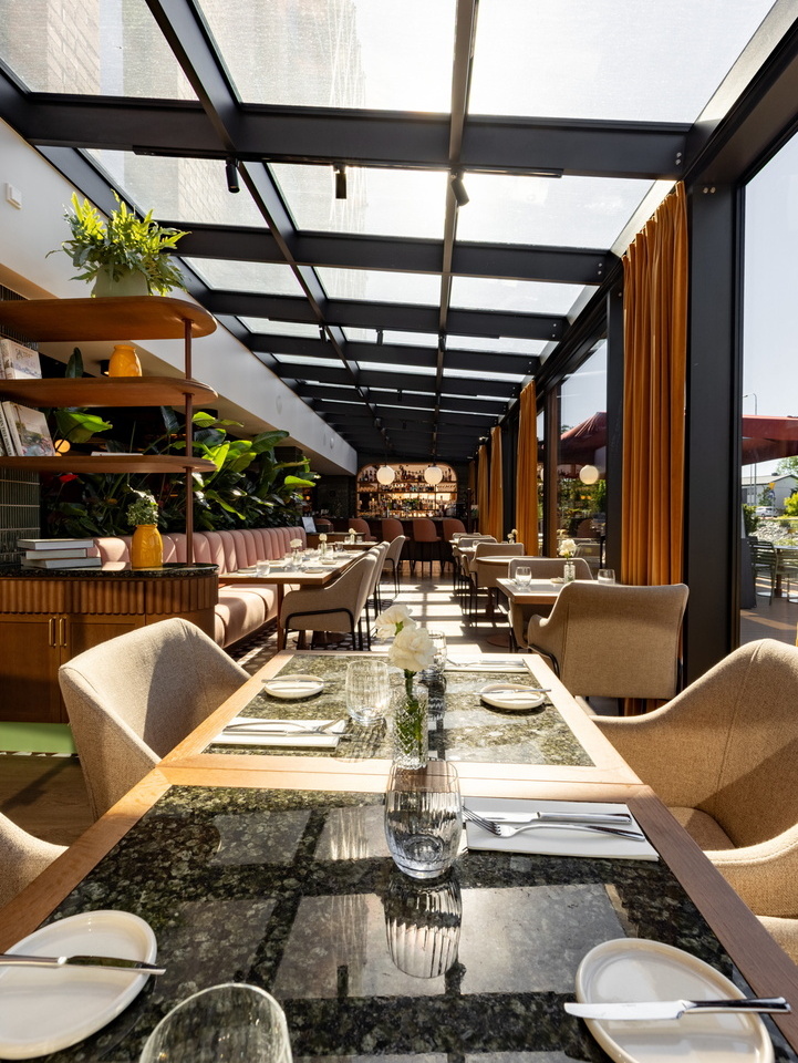 Szklana oranżeria-restauracja z naturalnym światłem, marmurowe stoliki, komfortowe fotele, rośliny i stylowy bar w tle