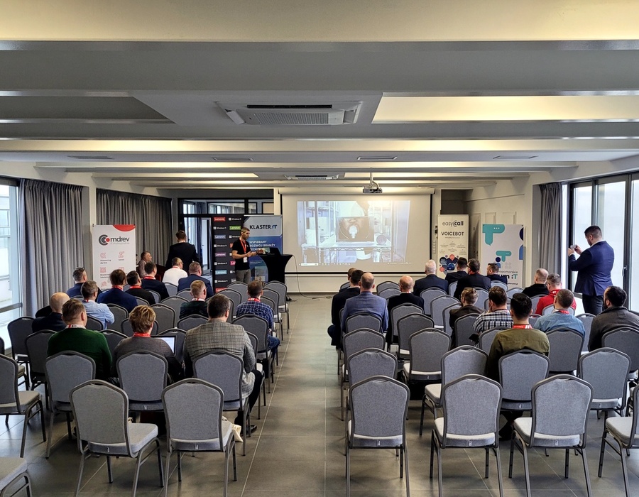 Modern conference room with rows of gray chairs, attendees watching a presentation projected on a screen.