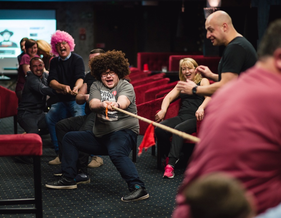 Hotel-Eventraum mit Gästen beim ausgelassenen Tauziehen zwischen roten Samtsesseln, Team-Building-Atmosphäre.