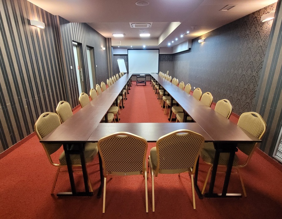 Konferenzraum in U‑Form mit langen Tischen, gepolsterten Stühlen, Projektorleinwand, Flipchart und warmer Beleuchtung