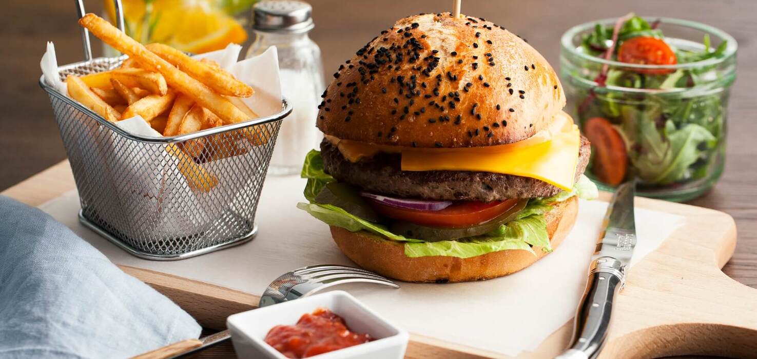 Burger z cheddarem na bułce z czarnym sezamem, sałata, pomidor i cebula; chrupiące frytki w koszyczku, sałatka i sos na drewnianej desce