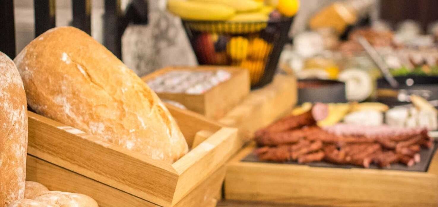 Świeże bochenki i pieczywo w drewnianych skrzynkach na bufecie śniadaniowym, w tle kosz z owocami i wędliny.
