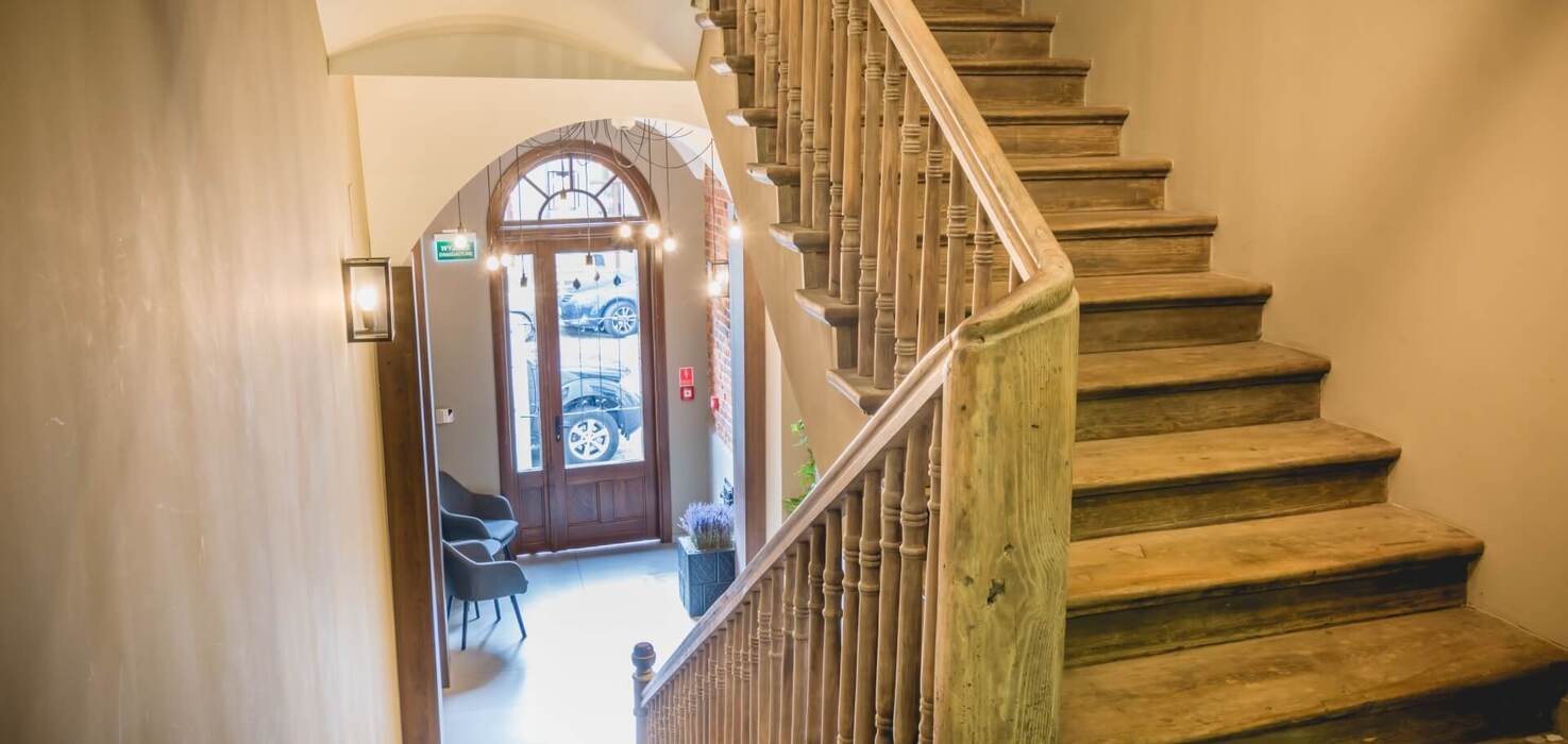 Drewniane schody z ozdobną balustradą, widok na przytulne wejście z fotelami, wiszącymi lampami i drzwiami z łukowym oknem.