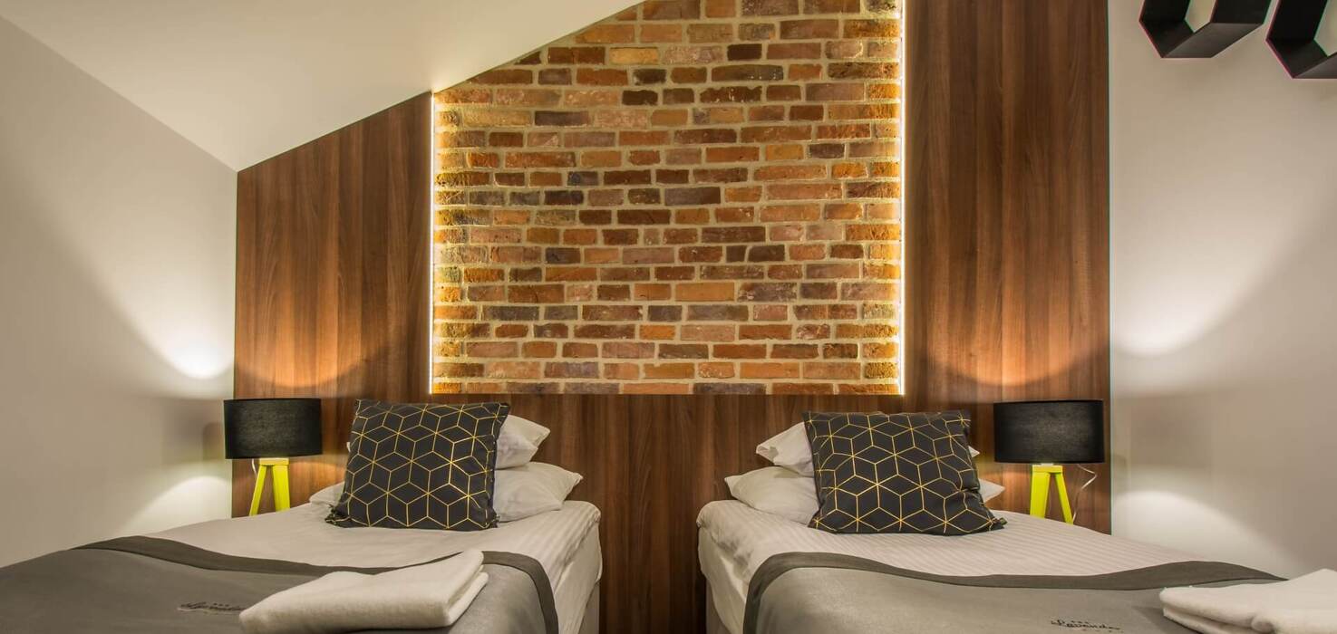 Pokój z dwoma pojedynczymi łóżkami, podświetlany ceglany panel nad wezgłowiem, drewniane panele i lampki nocne.