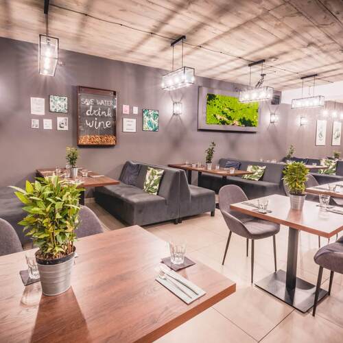 Nowoczesna restauracja hotelowa z drewnianym sufitem, wiszącymi lampami, szarymi kanapami, stolikami nakrytymi, roślinami i zielonym panelem na ścianie.