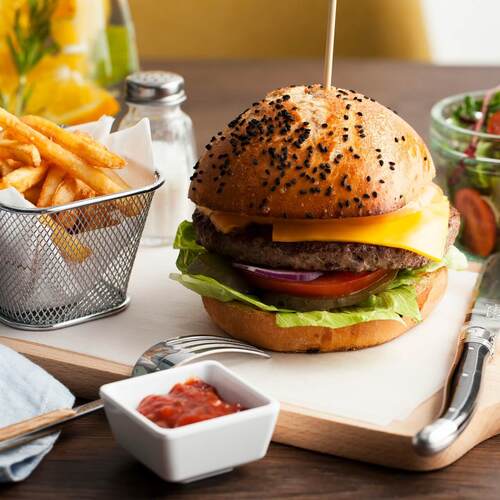 Burger z cheddarem na bułce z czarnym sezamem, sałata, pomidor i cebula; chrupiące frytki w koszyczku, sałatka i sos na drewnianej desce
