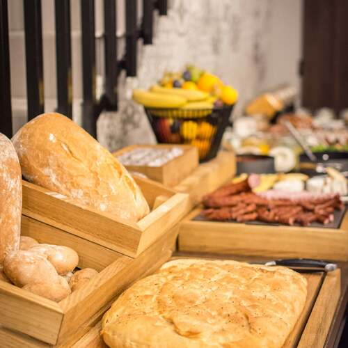 Świeże bochenki i pieczywo w drewnianych skrzynkach na bufecie śniadaniowym, w tle kosz z owocami i wędliny.