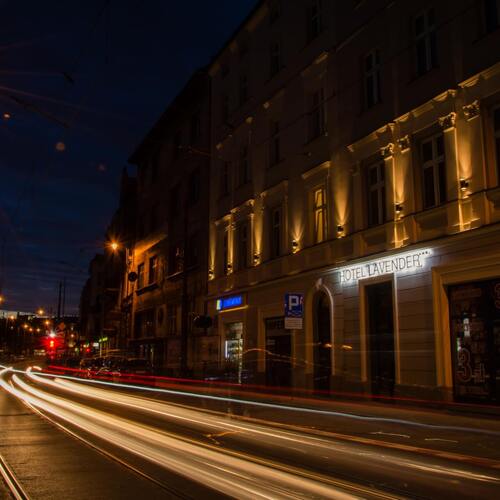 Oświetlona fasada Hotelu Lavender przy ruchliwej ulicy nocą, tramwajowe tory i świetlne smugi przejeżdżających samochodów