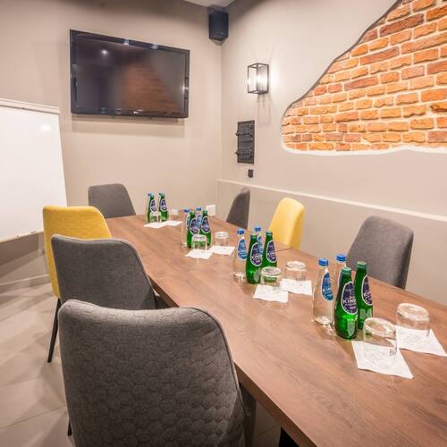Sala konferencyjna z drewnianym stołem, wygodnymi szarymi i żółtymi krzesłami, telewizorem, flipchartem i odkrytą cegłą.