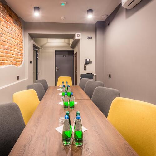 Sala konferencyjna z długim drewnianym stołem, szaro-żółtymi krzesłami, butelkami wody, ceglaną ścianą i klimatyzacją.