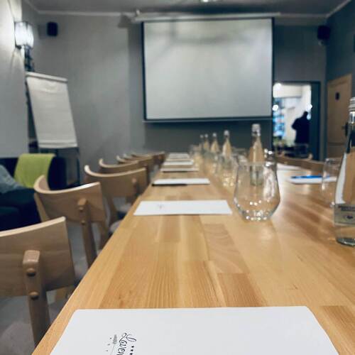 Sala konferencyjna hotelu z długim drewnianym stołem, krzesłami, ekranem rzutnika, butelkami wody i notesami