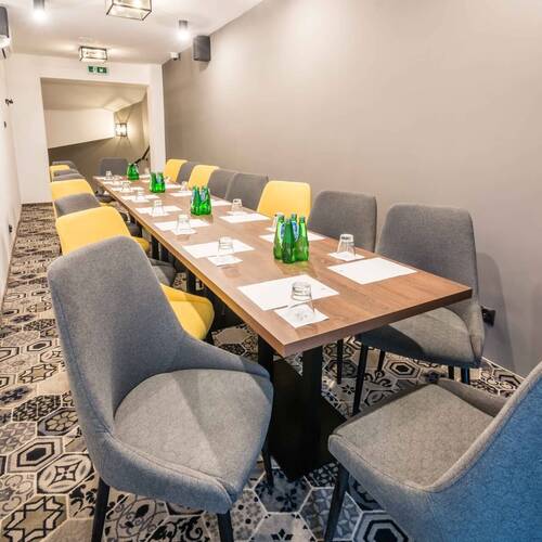 Sala konferencyjna z długim stołem, szaro‑żółtymi fotelami, butelkami wody, szklankami i notatkami; subtelne oświetlenie i dekoracyjny dywan.