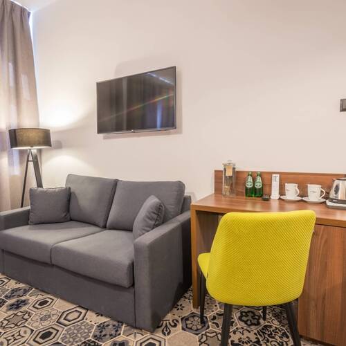 Przytulny hotelowy kąt z szarą sofą, lampą, ściennym telewizorem, biurkiem z czajnikiem, filiżankami i żółtym krzesłem