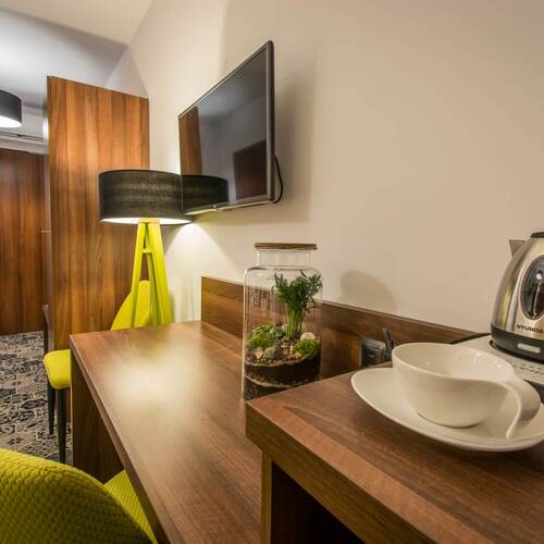 Biurko w pokoju hotelowym z czajnikiem, filiżankami, terrarium, wiszącym telewizorem, drewnianą zabudową i zielonym fotelem