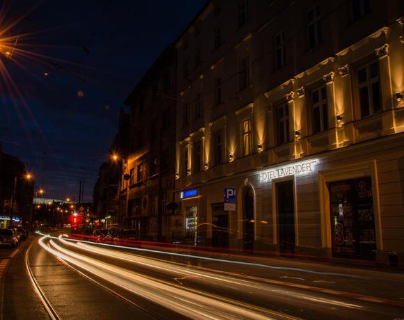 Oświetlona fasada Hotelu Lavender przy ruchliwej ulicy nocą, tramwajowe tory i świetlne smugi przejeżdżających samochodów