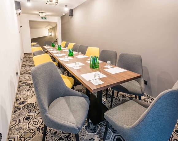 Sala konferencyjna z długim stołem, szaro‑żółtymi fotelami, butelkami wody, szklankami i notatkami; subtelne oświetlenie i dekoracyjny dywan.