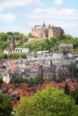 Historic castle atop lush green hill overlooking red-roofed town with medieval churches under blue sky