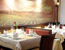 Elegant restaurant seating with white tablecloths, folded napkins, wine glasses, and a mural of vineyard scenery